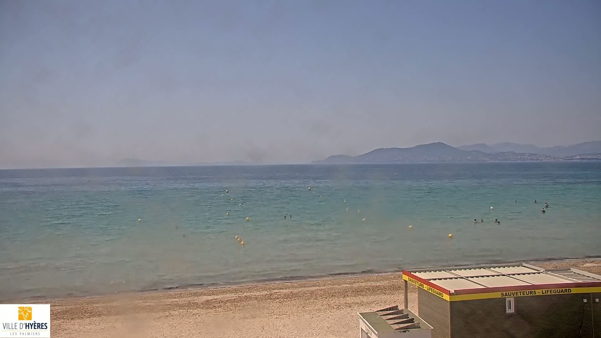 A sandy beach with calm, clear, teal water and a lifeguard station is visible, with hazy mountains in the background on a sunny day.