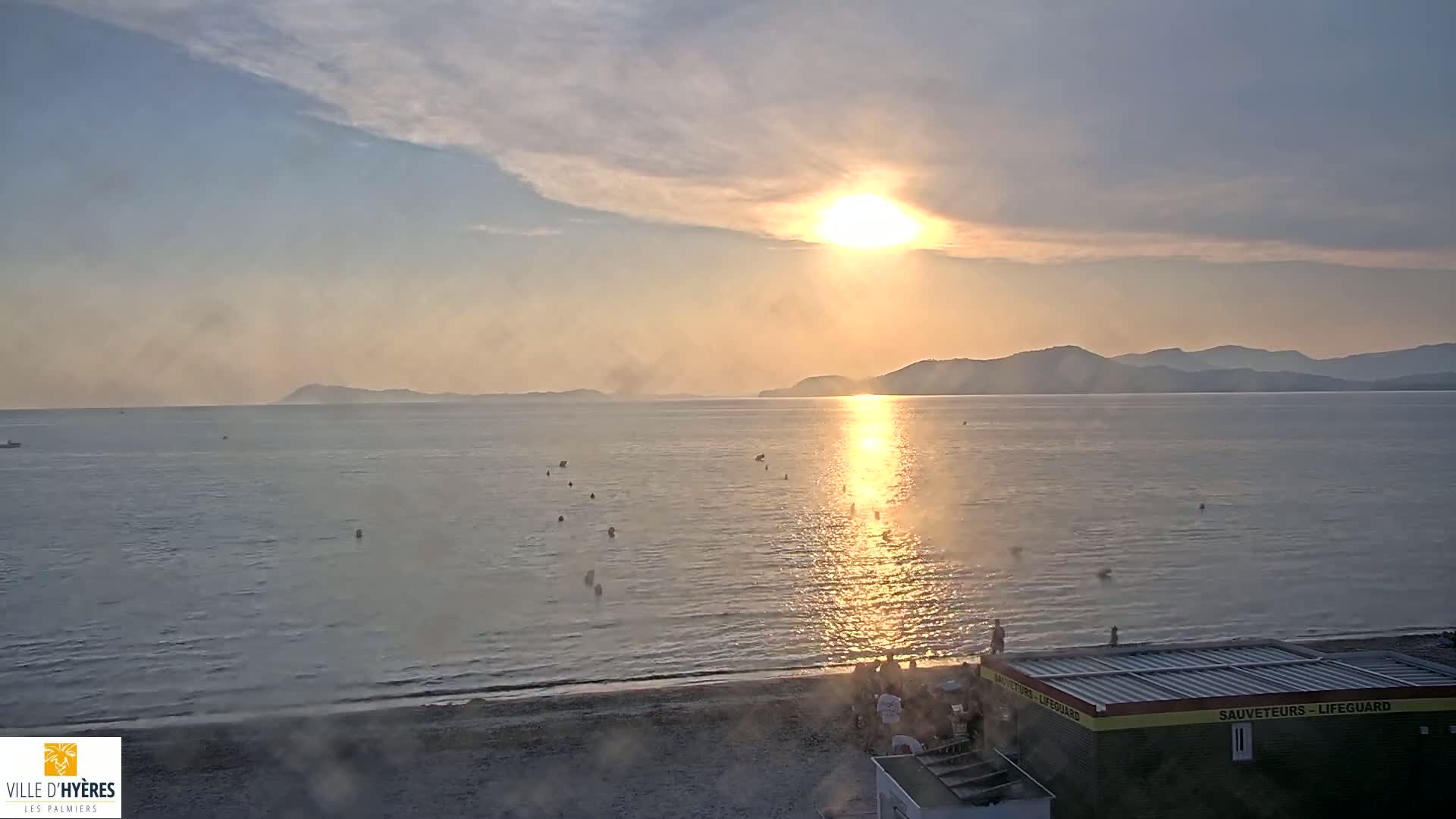 A hazy sunset over calm ocean water with a sandy beach and lifeguard station visible in the foreground.