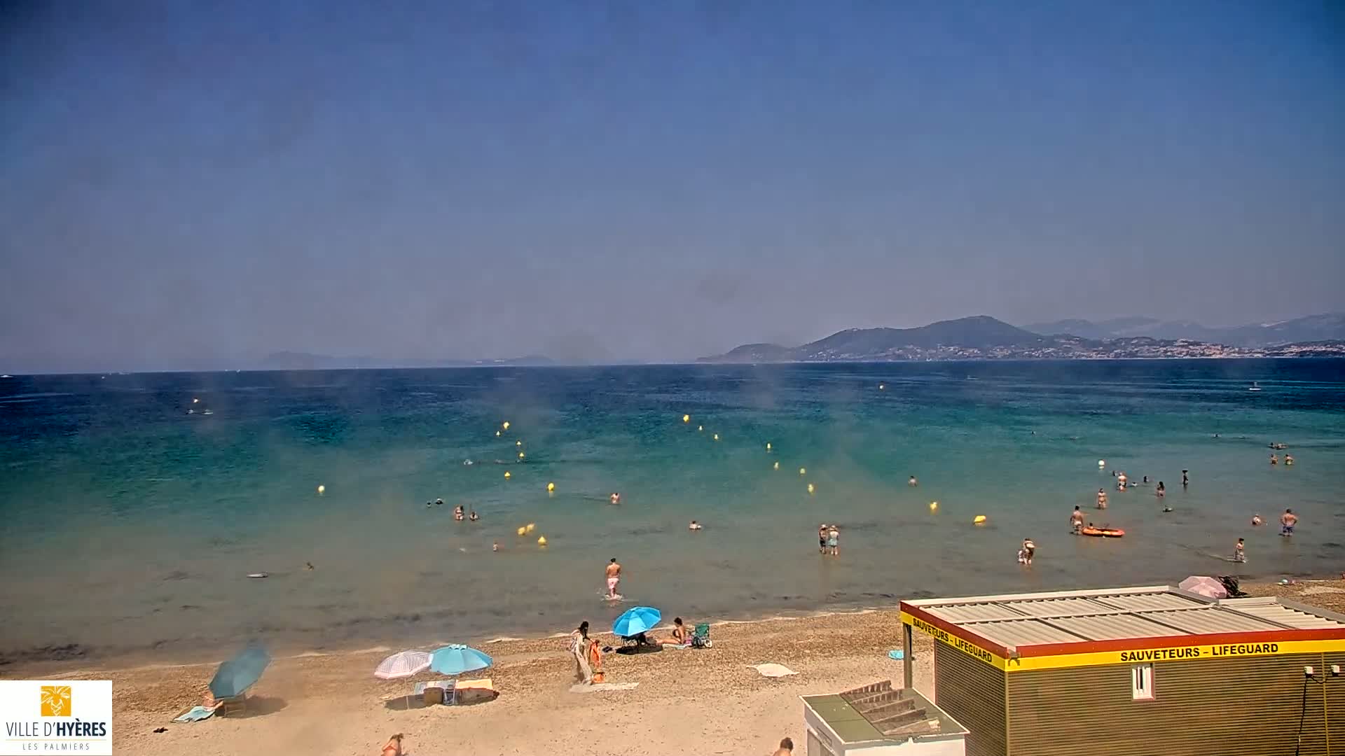 A hazy sunny day at a beach with people swimming and sunbathing, a lifeguard station, and mountains in the distance.