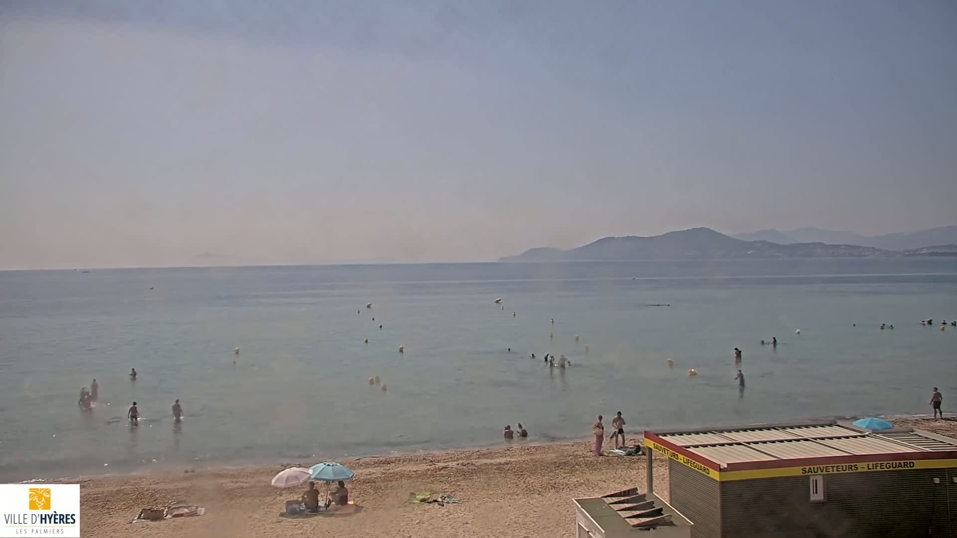 A hazy, sunny day at a beach with calm, clear water, people swimming and sunbathing, and a lifeguard station.