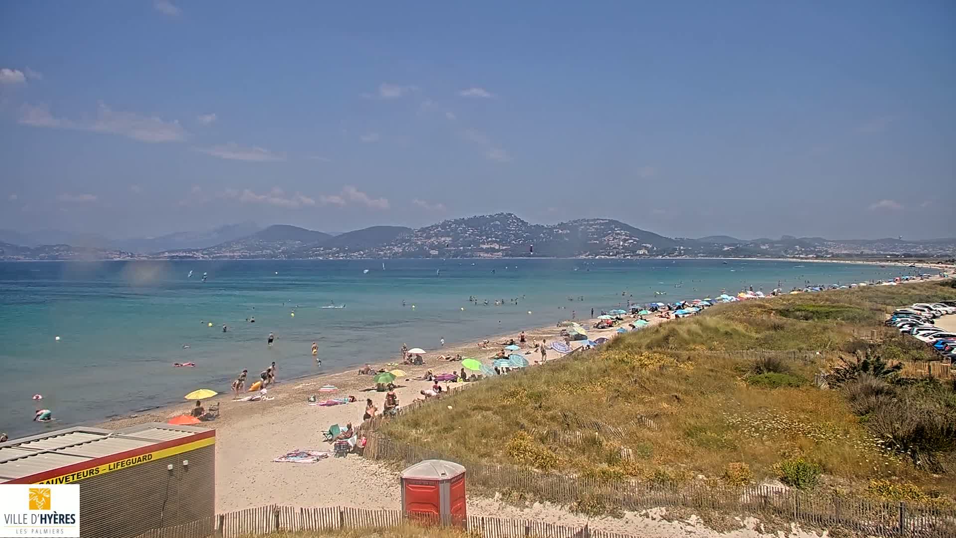 A sunny day at a beach with turquoise water, many people swimming and sunbathing under umbrellas, and distant mountains.