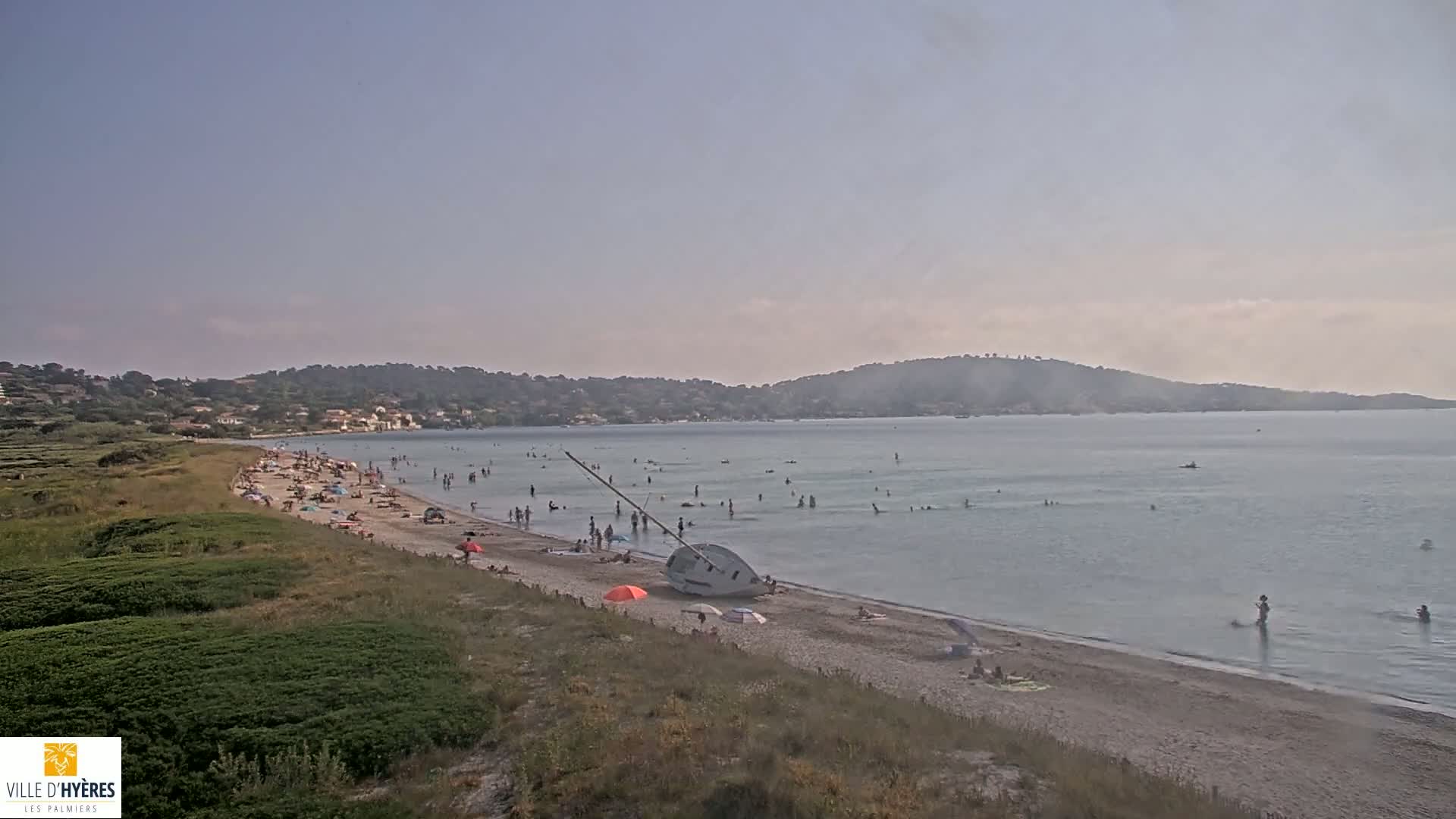 A sandy beach with many people swimming and sunbathing, a capsized sailboat on the shore, and a calm sea under a clear sky.