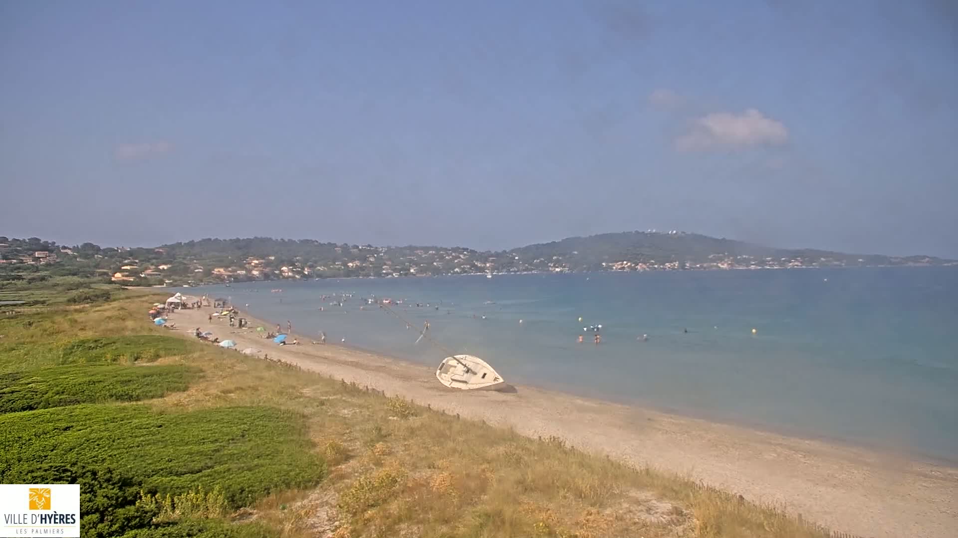 A sandy beach with scattered beachgoers, a capsized sailboat, and calm, clear water stretches toward a distant hilly coastline under a mostly clear sky.