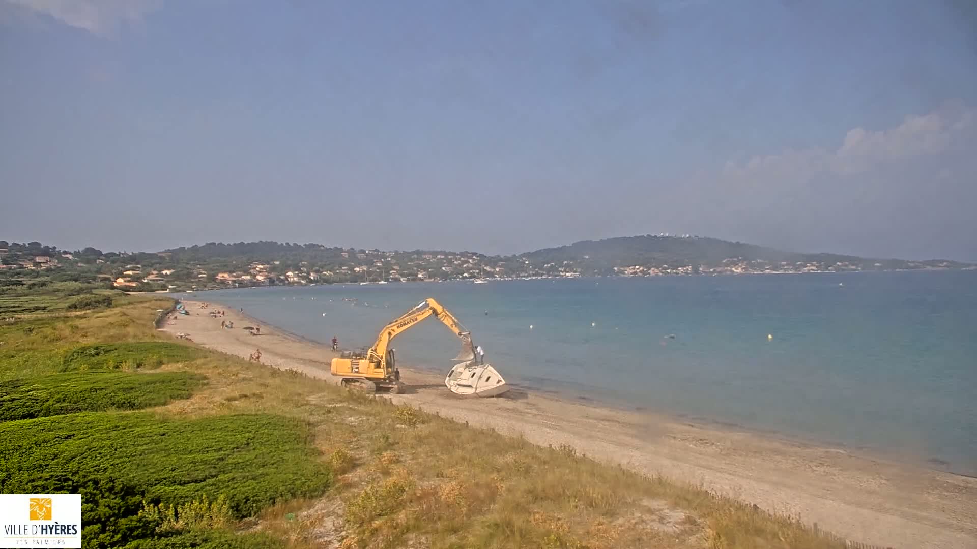 A yellow excavator moves a boat-like object on a sandy beach by calm, teal water under a clear, sunny sky, with a few people visible on the beach and a distant shoreline.