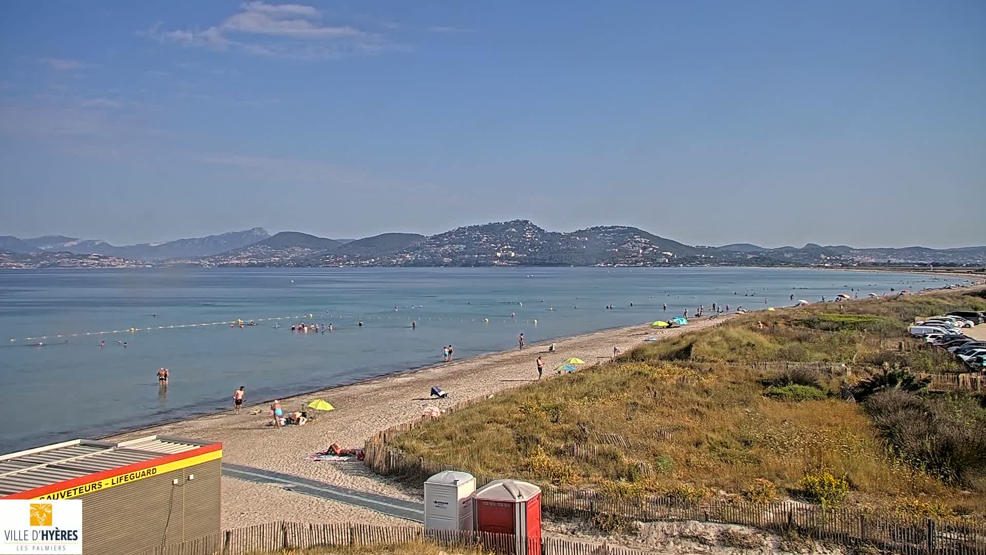 A sandy beach with bathers in shallow, calm water, backed by dry, grassy dunes and mountains in the distance under a clear, sunny sky.