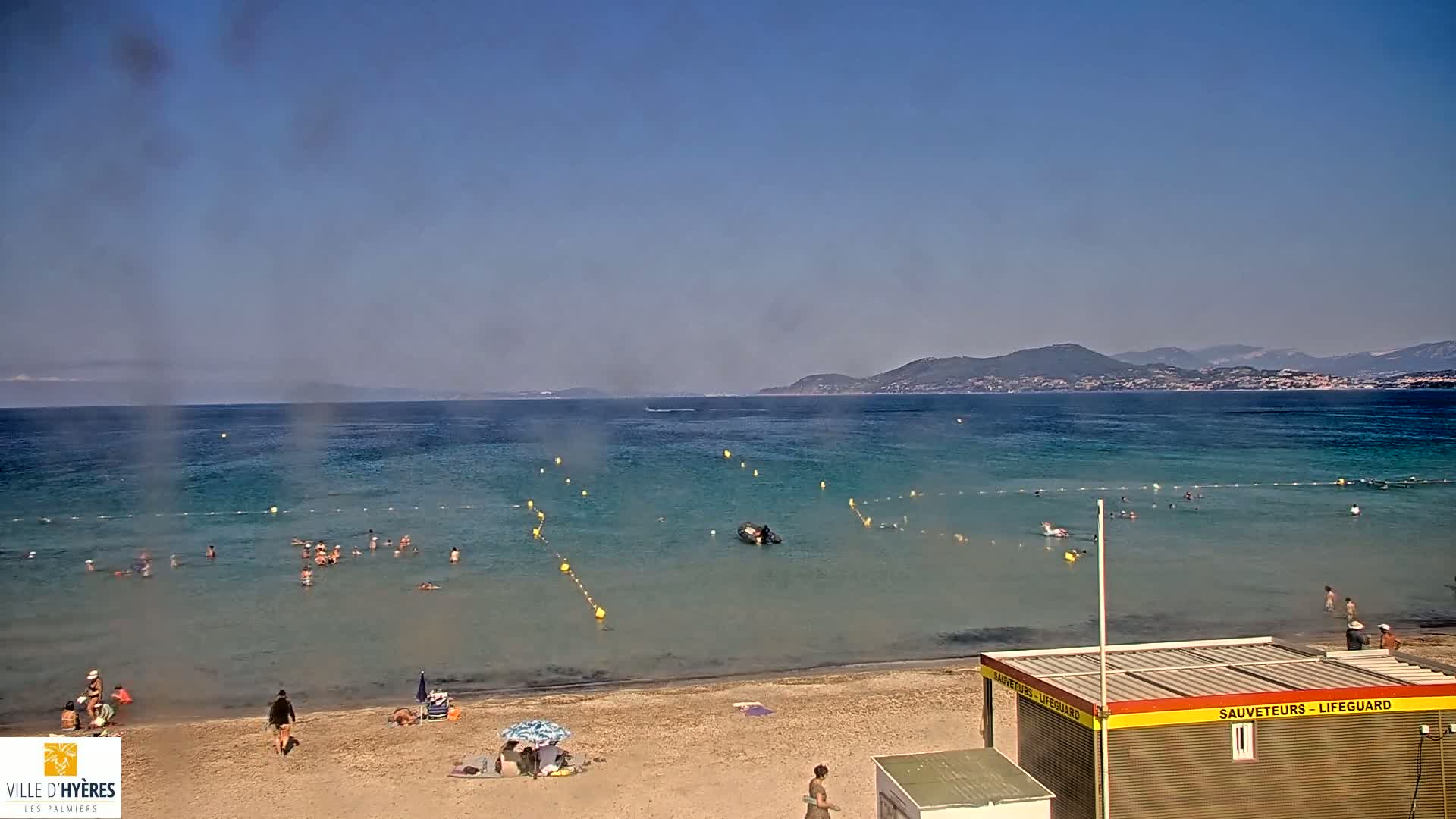 A sandy beach with people swimming and sunbathing in calm, clear water, under a hazy, sunny sky, with mountains in the distance.