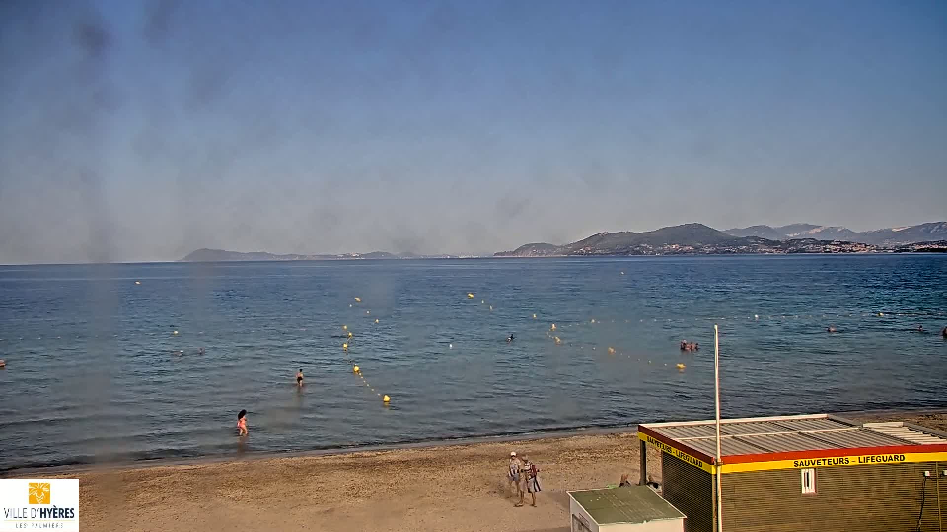 A sandy beach with calm, clear water, swimmers, and a lifeguard station is visible, with mountains in the background on a hazy day.
