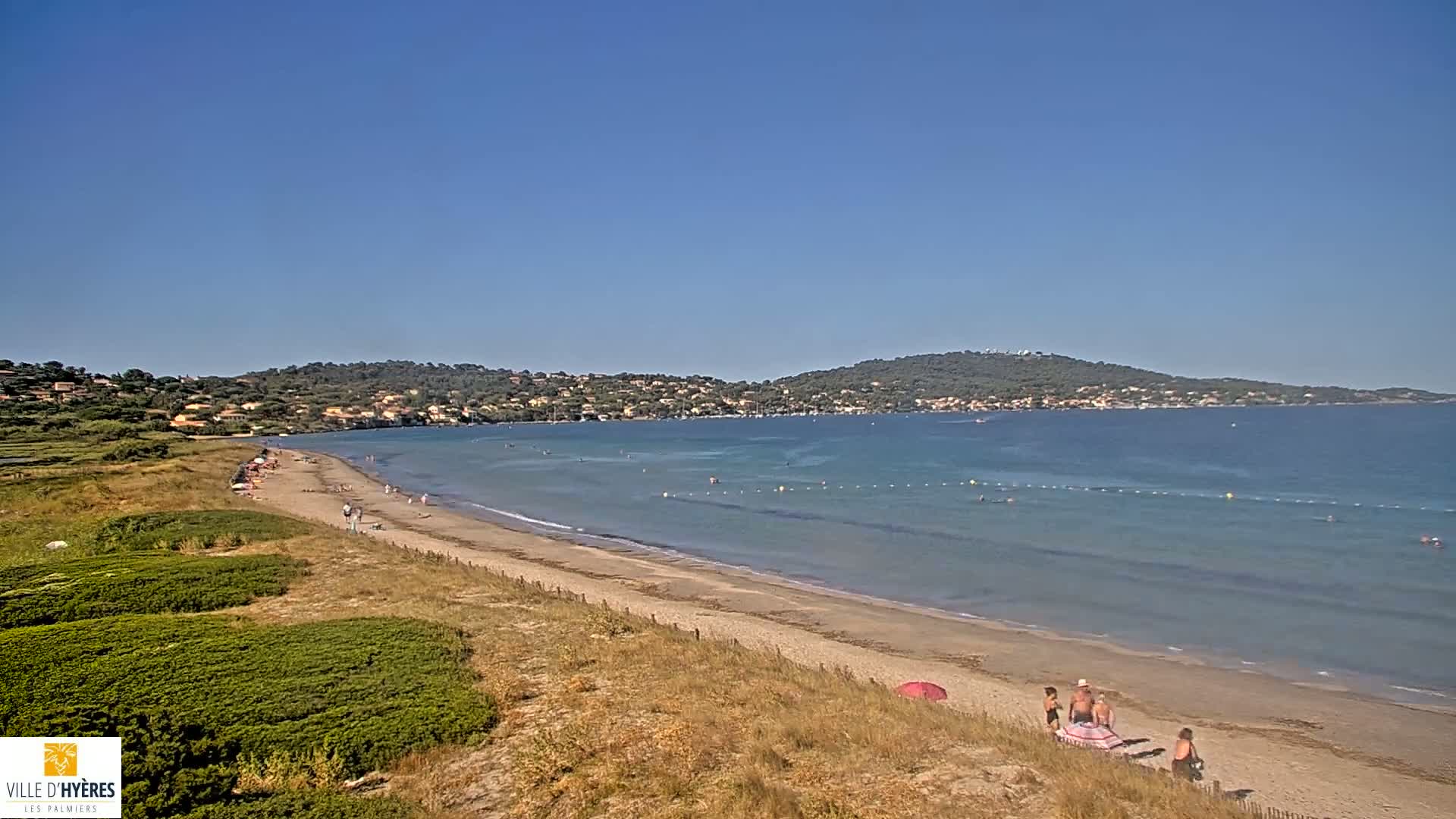 A sandy beach with calm, clear water stretches out to a distant shoreline under a clear blue sky, with a few people visible on the beach.