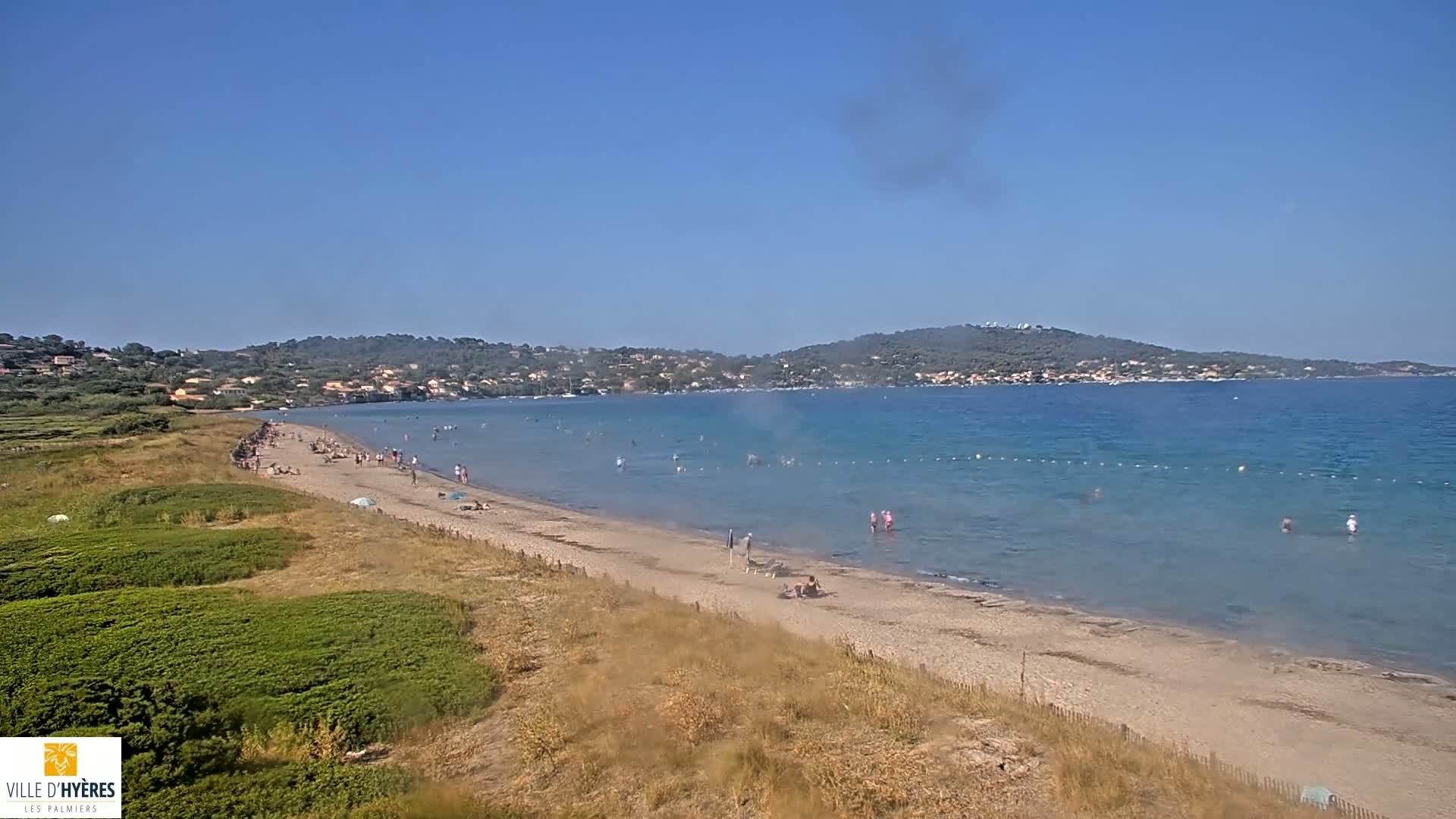 A sandy beach with scattered people on it and in the calm, clear water, extends to a calm bay and hills in the distance under a clear blue sky.