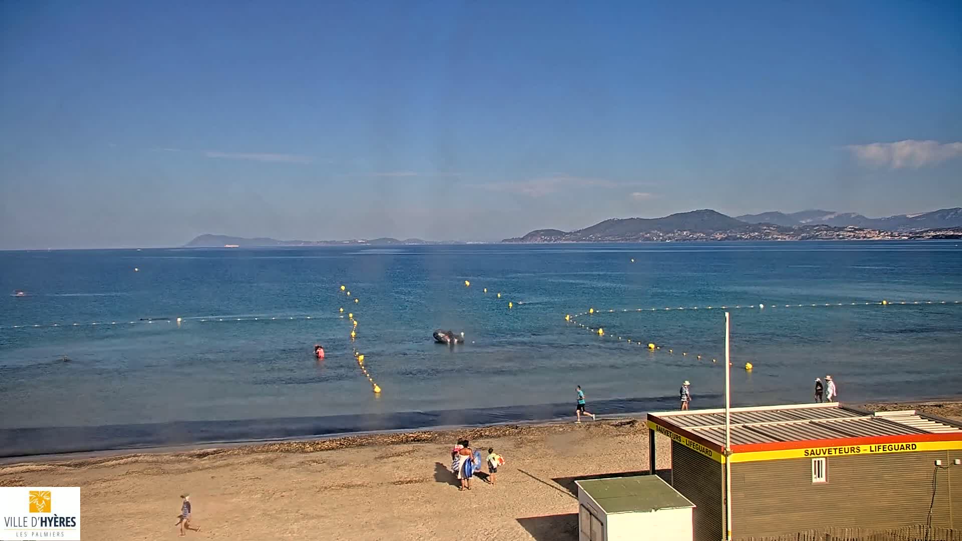 A sandy beach with several people on it and in the shallow, calm, blue water, is bordered by a lifeguard station, with mountains visible in the distance under a clear blue sky.