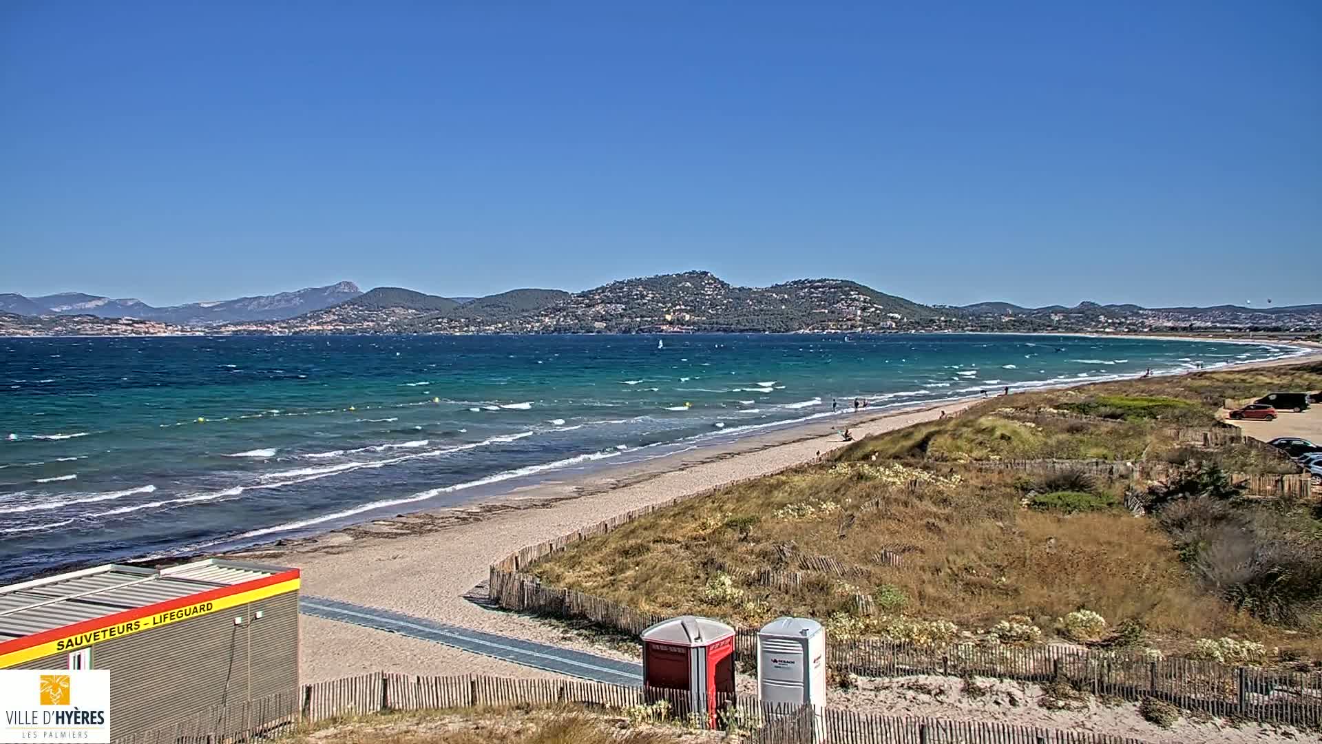 A sandy beach with small waves under a clear blue sky stretches toward a distant mountainous coastline.