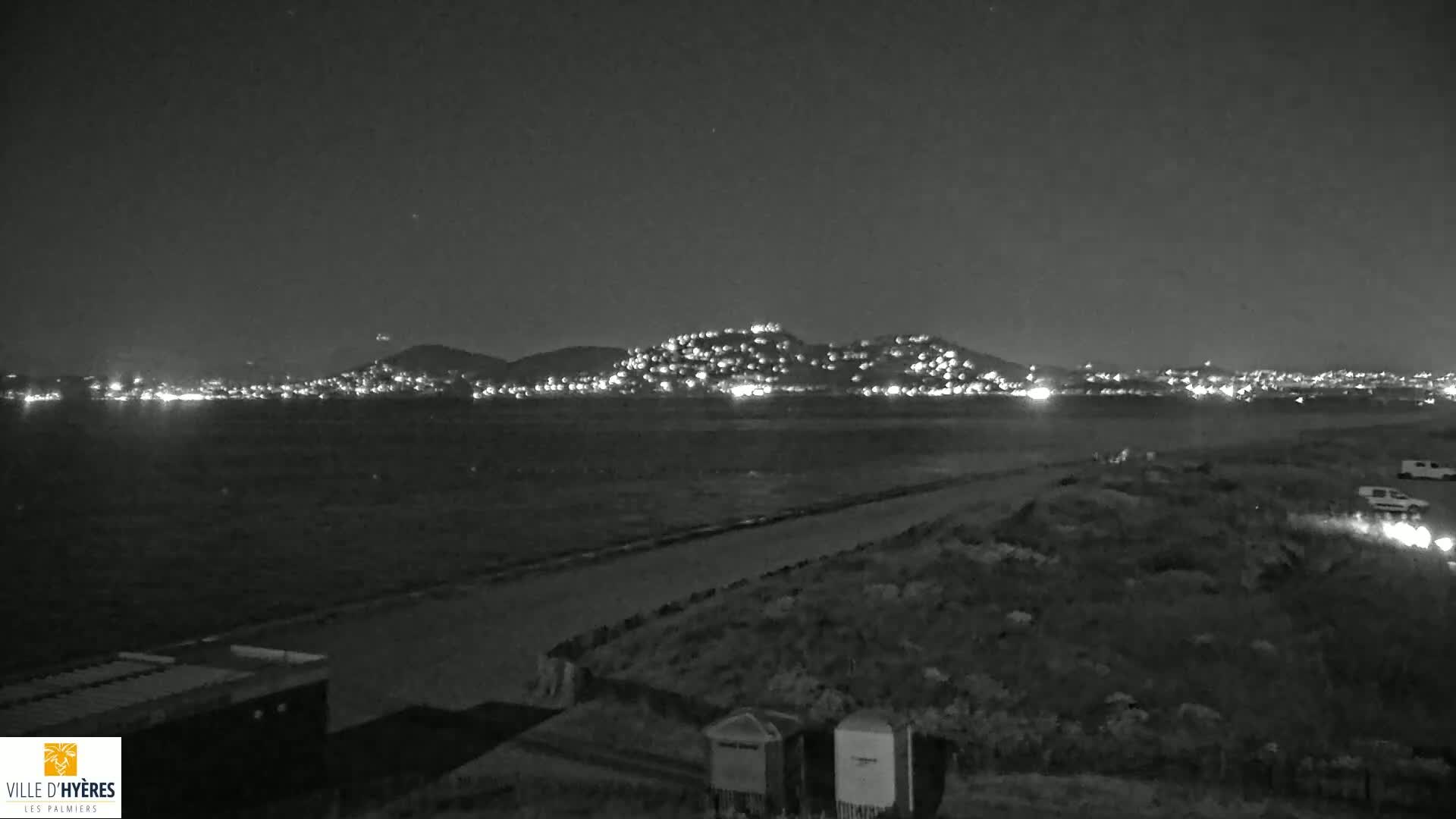 A nighttime view shows a body of water with a hill dotted with lights in the background under a clear, dark sky.