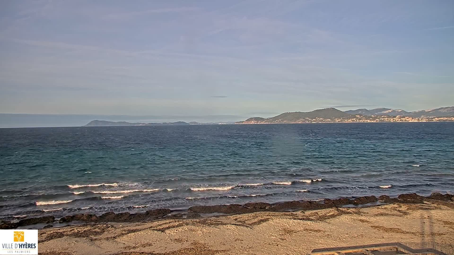 A coastal landscape shows a sandy beach with seaweed and scattered rocks in the foreground, leading to a blue-green sea with gentle waves, all set against a distant mountainous coastline dotted with towns under a clear, bright sky.