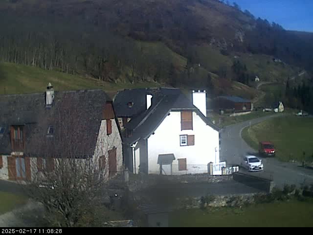 Two houses sit on a hillside overlooking a valley under a clear, sunny sky, with a couple of cars parked on a road nearby.