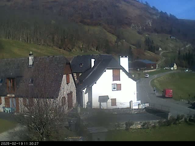 A sunny day in a rural valley shows several stone buildings, a winding road, and a person walking near a red truck.