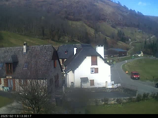 A sunny day reveals a small village nestled in a valley, with several stone buildings and a winding road, surrounded by brown and green hillsides.