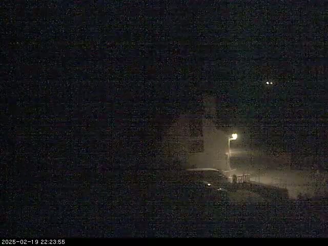 A dimly lit nighttime scene shows a vehicle parked near a building under a dark, possibly snowy sky.