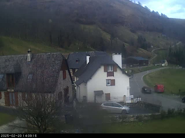 A cluster of houses nestled in a valley on an overcast day, with a car parked in front of one of the houses and another car and truck driving on a nearby road.