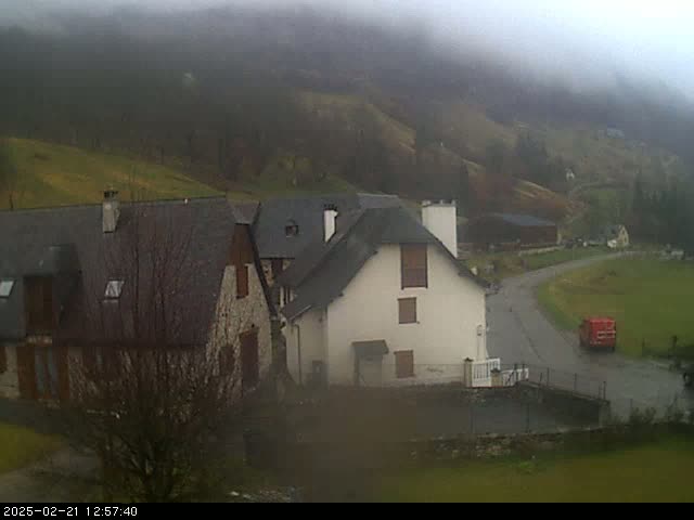 A foggy, overcast day reveals a cluster of stone houses nestled in a valley, with a red vehicle visible on a road below.
