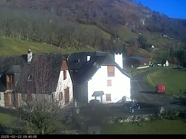 Two houses sit on a hillside overlooking a paved road and a valley under a clear, sunny sky.