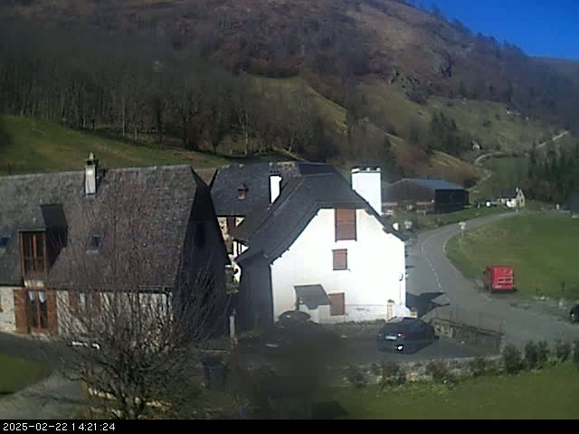 A sunny day reveals a cluster of stone buildings nestled in a valley, bordered by a gently sloping hillside covered in sparse trees.