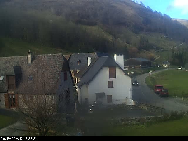 A small village nestled in a valley on a sunny day, with several stone buildings and a few vehicles on a winding road.