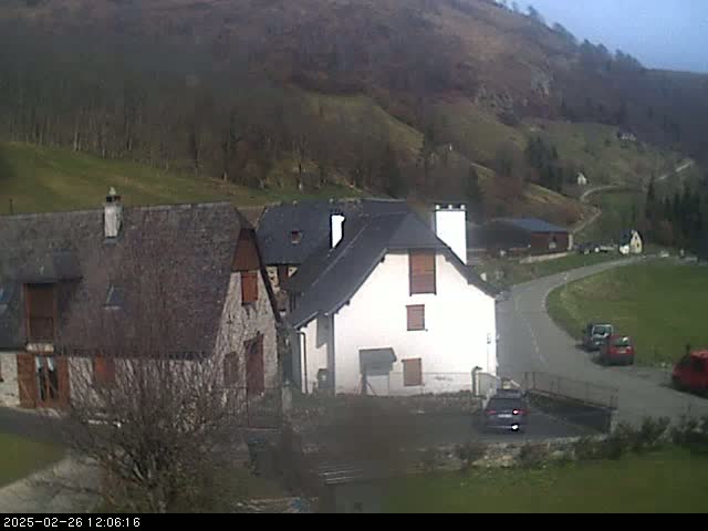 A small village nestled in a valley on a partly cloudy day, with several stone buildings and a few cars parked on a winding road.