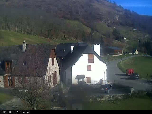 A sunny day reveals several stone buildings nestled in a valley surrounded by hillsides with sparse trees.