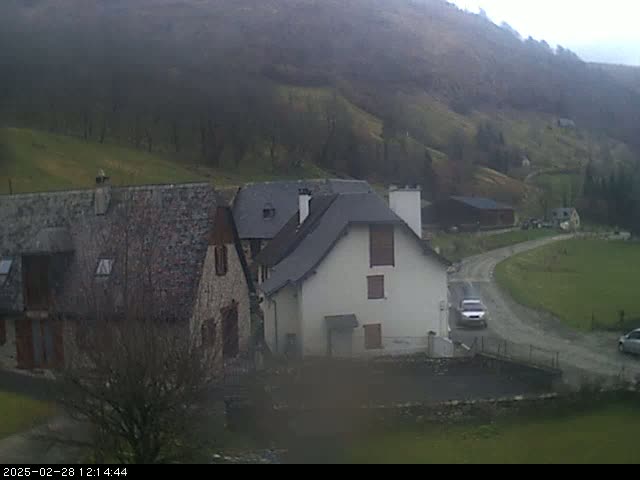 A cloudy day reveals a rural scene of stone buildings nestled in a valley, with a gravel road curving through a grassy area.