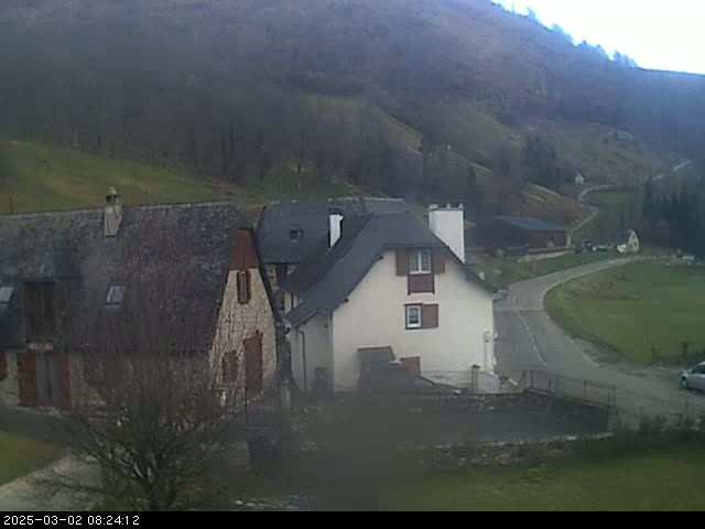 A white house and a stone building sit on a hillside overlooking a road that curves through a valley under a cloudy sky.