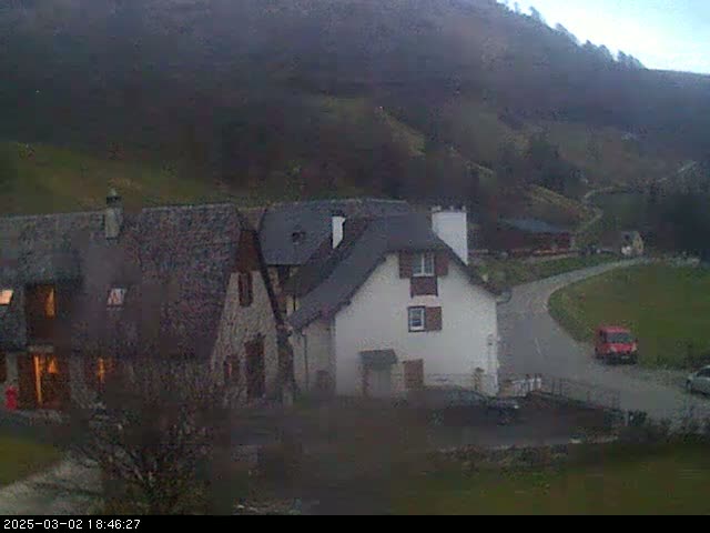 A small village nestled in a valley on an overcast day, with a few buildings and a red van visible on a winding road.
