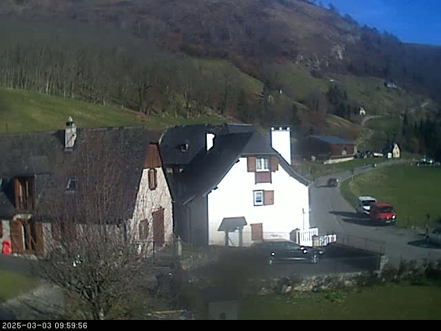 A sunny day reveals a hillside village with several buildings, including a white farmhouse and a stone house, nestled among trees and a gently sloping green field.