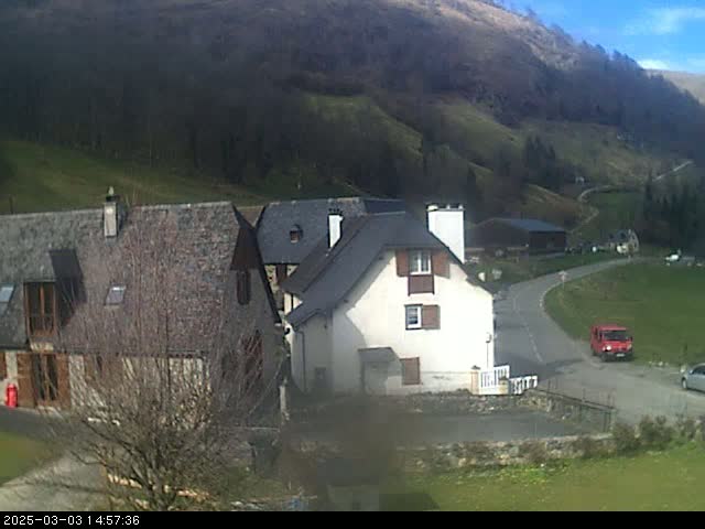 A small village nestled in a valley on a sunny day, with several stone buildings and a red van parked on a road.