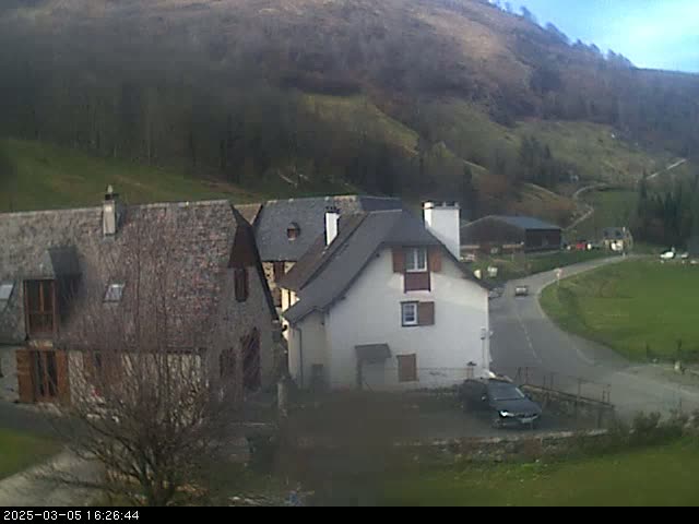 A small village nestled in a valley on a partly cloudy day, with several stone buildings and a car parked on a road curving through the scene.
