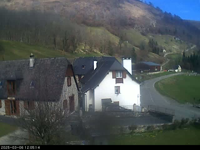 A sunny day reveals a rural scene with stone buildings nestled in a valley, surrounded by a gently sloping hillside dotted with trees.
