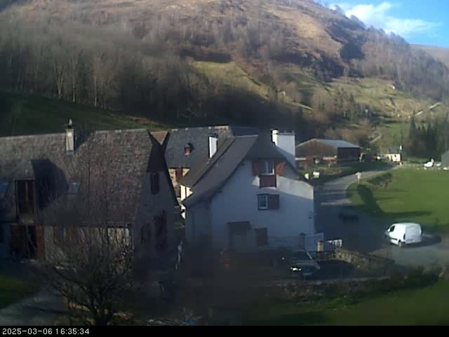 A sunny day reveals a cluster of houses nestled in a valley, with a hill covered in trees and sparse vegetation in the background; a white van is parked on a road near the houses.
