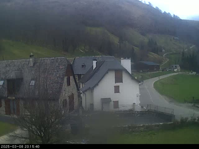 A rural scene on an overcast day shows several stone buildings nestled in a valley surrounded by rolling green hills.