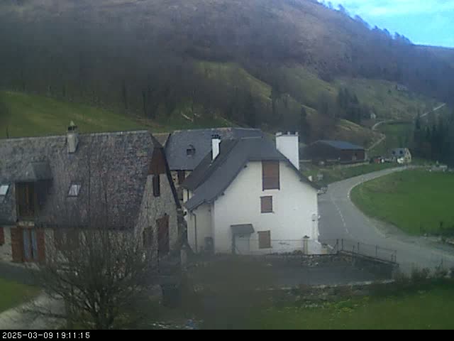 Stone houses and farm buildings sit on a hillside overlooking a curving road, under a mostly cloudy sky.