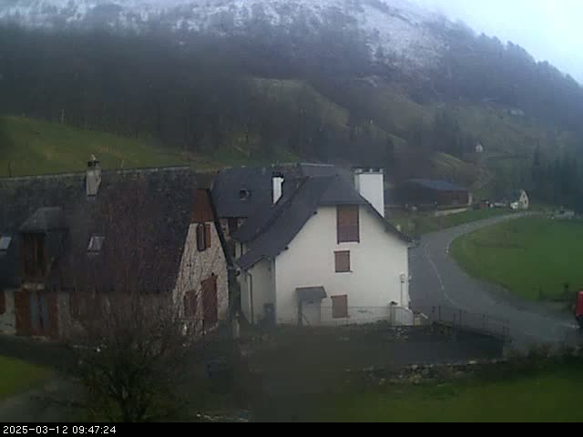 A misty, overcast day reveals a snow-dusted mountain overlooking a rural village scene with stone houses and a curved road.