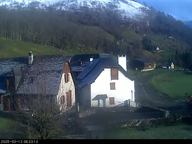 A snow-dusted mountain overlooks a small village of stone and slate-roofed houses on a partly sunny day.