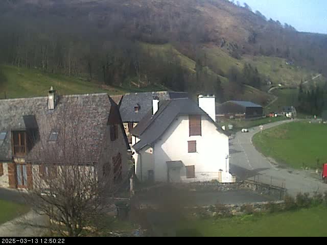 A cluster of stone buildings sits on a hillside overlooking a road curving through a valley on a sunny day.