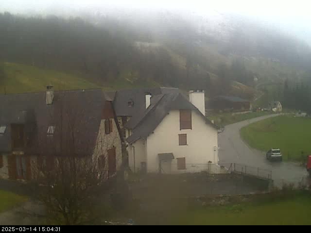 A foggy view of a small village nestled in a valley, with several stone buildings and a car parked on a curved road.