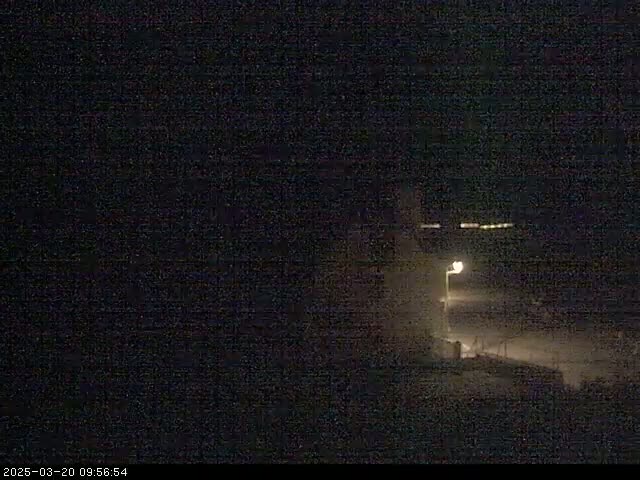 A dimly lit, dark scene at night shows a building and a streetlamp, possibly near the coast.