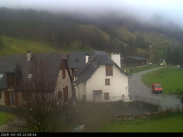 A foggy view of a small village nestled in a valley, with stone buildings and a red van on a road.