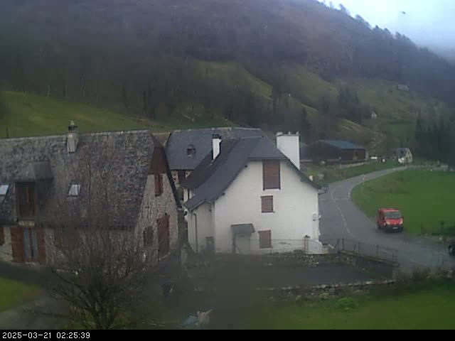 Stone buildings sit on a hillside overlooking a valley under an overcast sky, with a red van visible on a nearby road.