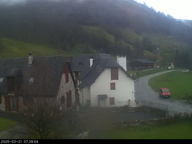 A misty, overcast day reveals a rural scene with stone buildings, a paved road, and a red van parked near a small bridge in a valley surrounded by hills.