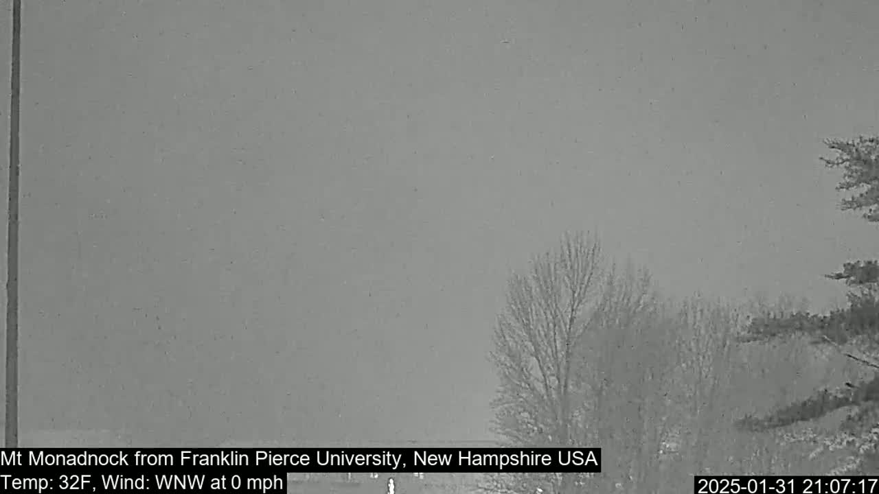 A gray, overcast sky hangs over a snow-covered landscape with bare trees and a road.