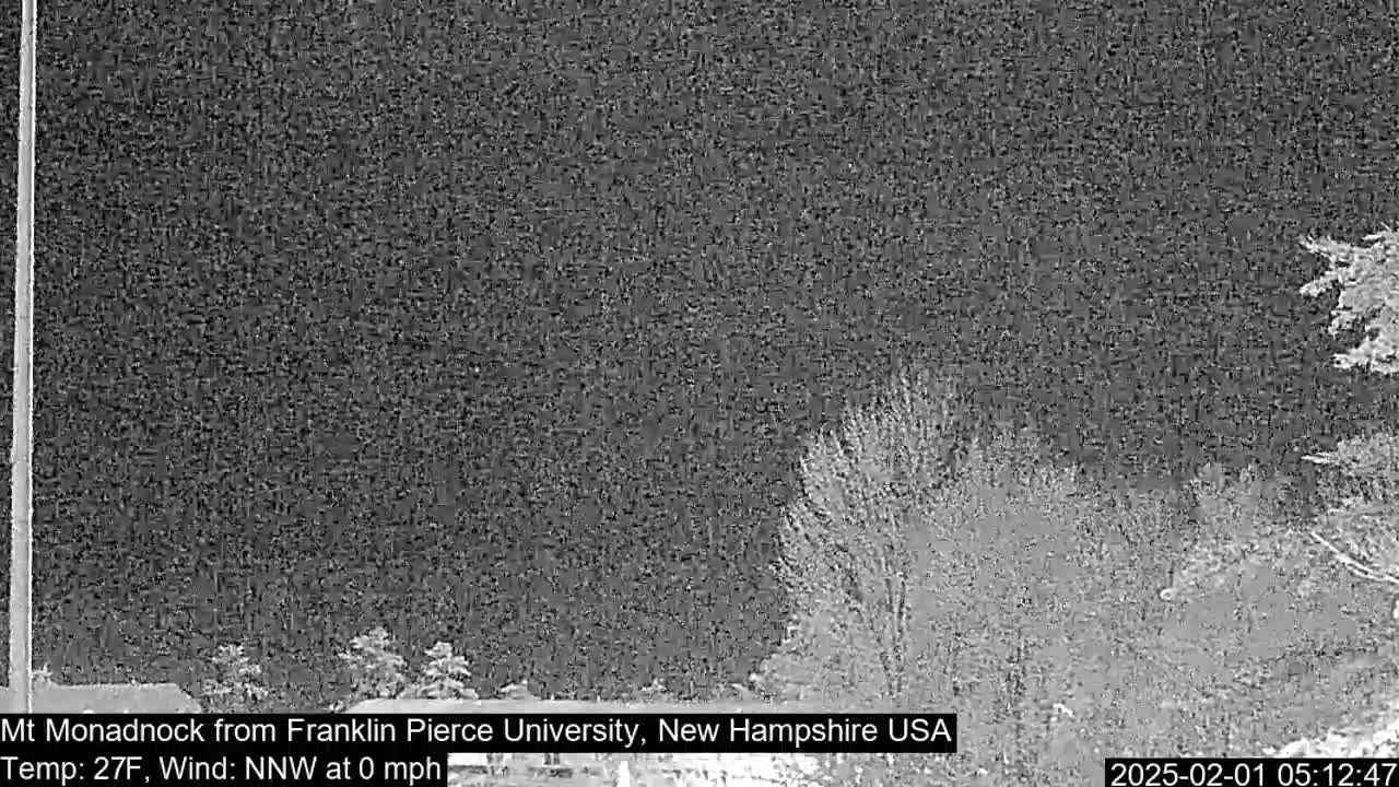 A nighttime, low-light image shows a dark, mostly clear sky over a snow-covered landscape with some leafless trees and shrubs.