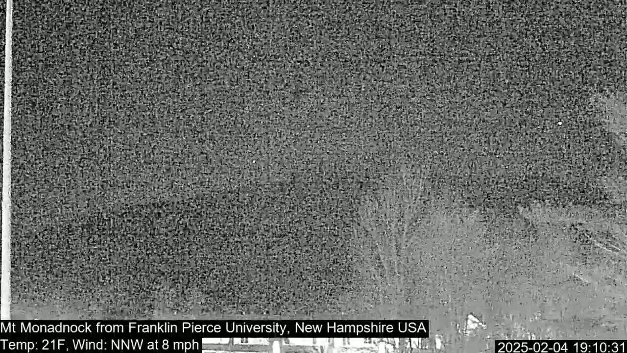 A nighttime, low-resolution image shows a dark, snowy landscape under a cloudy sky with a north-northwest wind at eight miles per hour and a temperature of 21 degrees Fahrenheit.