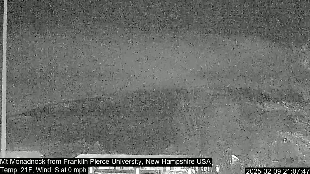 A nighttime, low-light view of a distant, snow-covered mountain range under a cloudy sky with calm winds and a temperature of 21°F.